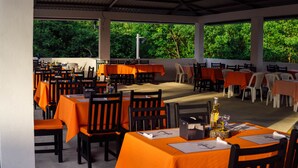 An outdoor dining area with tables covered in orange tablecloths, surrounded by chairs, and large windows offering a view of greenery. - Hotel Majahual Resort  (La Libertad)