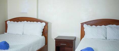 Deux lits simples avec des têtes de lit en bois et du linge de lit blanc, une petite table de chevet en bois placée entre les deux, et une serviette bleue sur chaque lit.