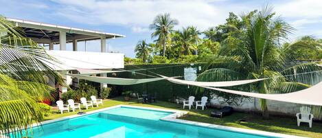A swimming pool with white chairs, surrounded by palm trees and a covered seating area.