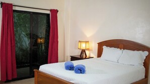 A neatly made bed with white bedding and blue towels, a wooden headboard, a nightstand with a lamp, and red curtains. - Hotel Majahual Resort  (La Libertad)