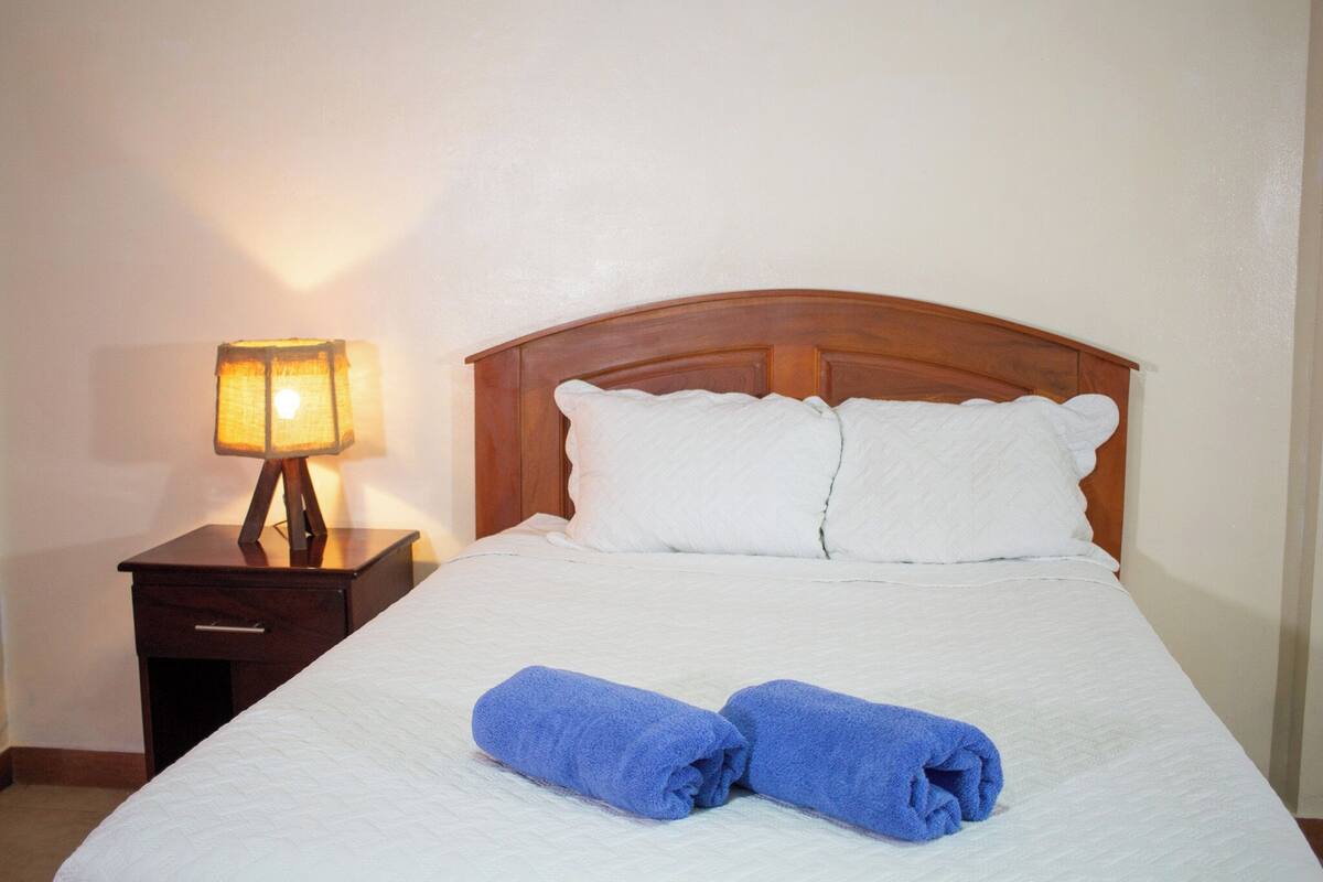 A neatly made bed with white linens and two rolled blue towels, a wooden headboard, and a bedside lamp on a wooden nightstand.