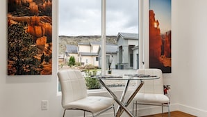 A modern dining area with a glass table and two white chairs, overlooking a residential neighborhood with houses and a rocky landscape.