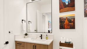 A modern bathroom with a white vanity, a large mirror, and a wooden cabinet. There are two framed landscape paintings on the wall.