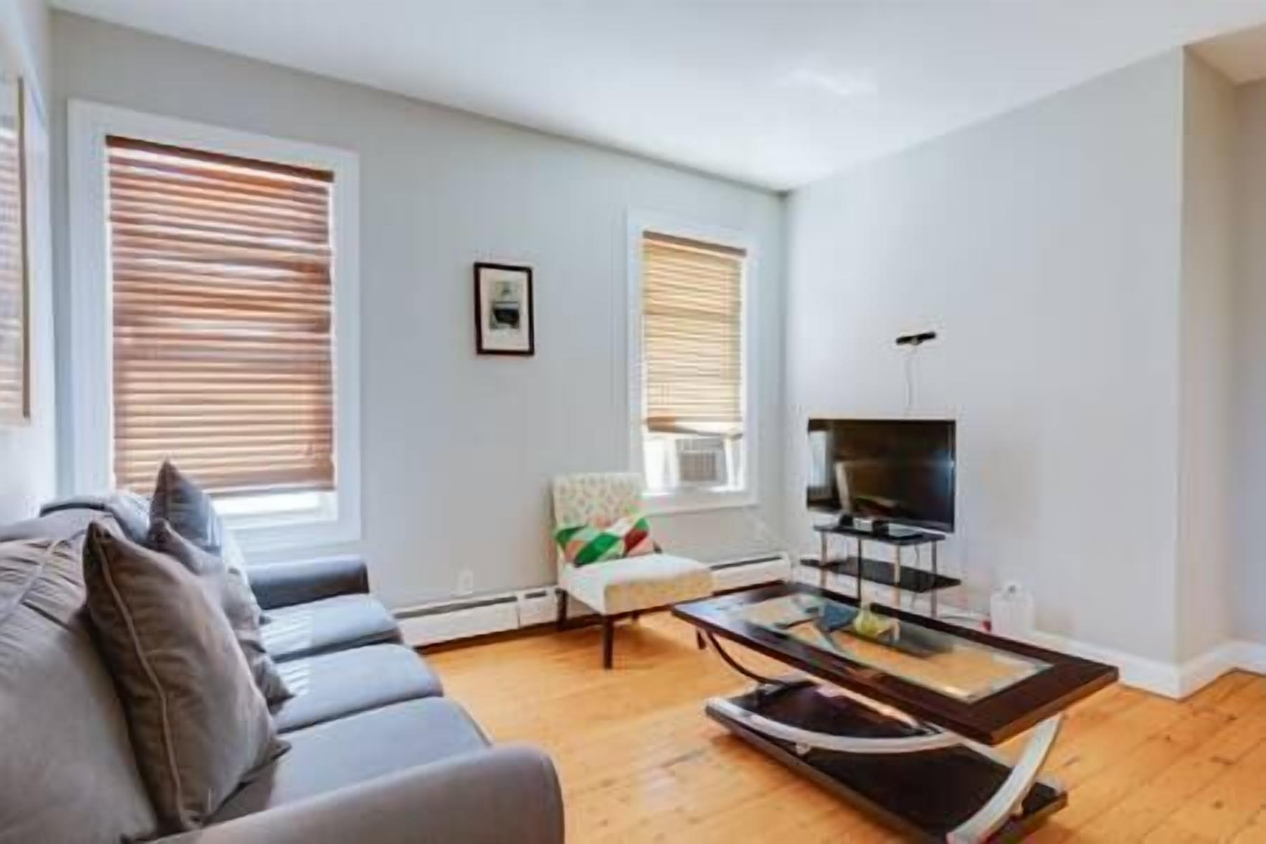 A living room with a grey sofa, a glass coffee table, a TV on a stand, and two windows with blinds.