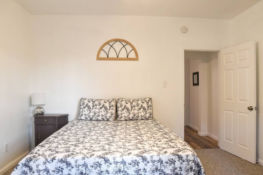 A bedroom with a floral bedspread, a nightstand with a lamp, a wooden-framed window, and a white door.