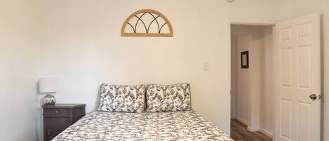 A bedroom with a floral bedspread, a nightstand with a lamp, a wooden-framed window, and a white door.