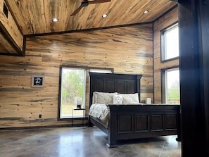 A bedroom with a wooden ceiling, a large window, and a bed with a dark headboard.