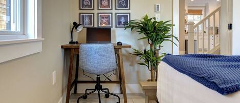 A bedroom with a bed, a desk with a chair, a potted plant, and framed artwork on the wall.