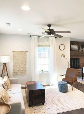 Relax in this inviting living space with a sofa, coffee table, and side table, surrounded by a patterned rug and large window with white curtains.