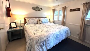 Un dormitorio con una cama, una guitarra, una mesita de noche, una lámpara, un ventilador de techo y una ventana con cortinas.