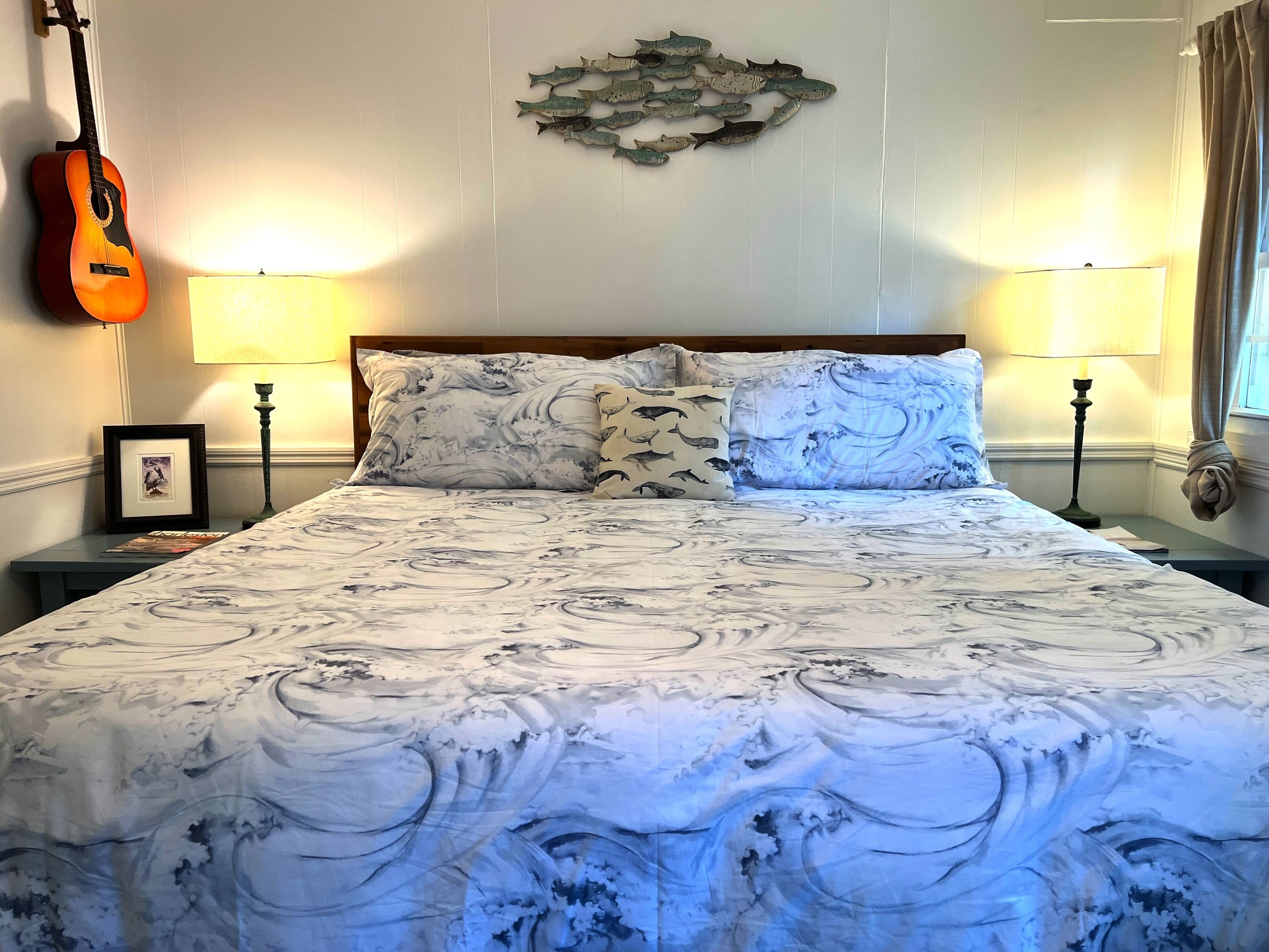 Una cama con colcha de diseño marmolado, dos almohadas con motivo de olas, una guitarra en la pared y dos lámparas fijadas a la pared.