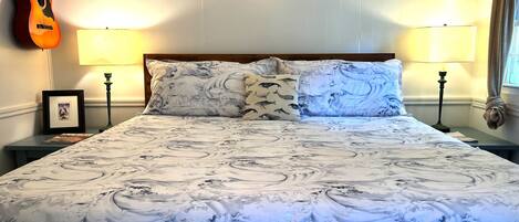 Una cama con colcha de diseño marmolado, dos almohadas con motivo de olas, una guitarra en la pared y dos lámparas fijadas a la pared.