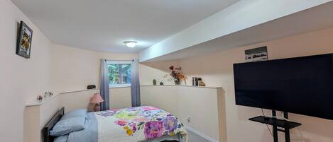 Une chambre à coucher comprenant un lit, un téléviseur, une fenêtre avec des rideaux, une étagère avec des objets décoratifs et un luminaire au plafond.