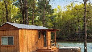 A wooden cabin with a metal roof, a small porch, and a dock extending into a lake surrounded by trees.