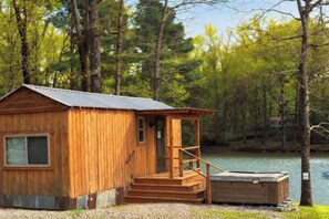 A wooden cabin with a metal roof, a small porch, and a dock extending into a lake surrounded by trees.