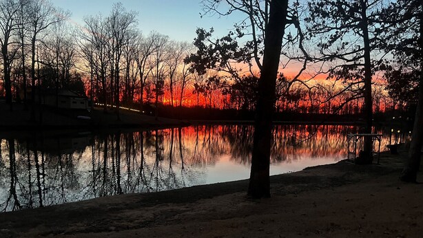Een bad in een rustgevende natuurlijke omgeving met een zonsondergangreflectie op het water, omringd door bomen en een gebouw in de verte.