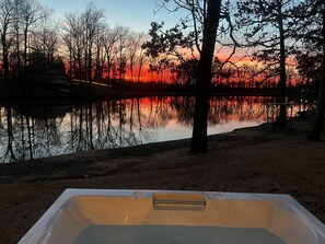 Une baignoire installée dans un cadre extérieur paisible, où se reflète le coucher de soleil sur l’eau, entourée d’arbres et d’un bâtiment situé au loin.