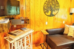 A room with a wooden wall, a mounted television, a wooden table with books, a black leather sofa, and a decorative tree wall art. - Lakefront Tiny Cabin Near Memphis + Hot Tub (Oakland)