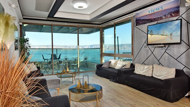 A modern living room with a large glass wall, a black sofa, a coffee table, and a TV mounted on the wall. - luxury suites (Tiberias)
