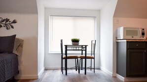 Um quarto pequeno e claro com janela, uma mesa com duas cadeiras, micro-ondas e um armário.