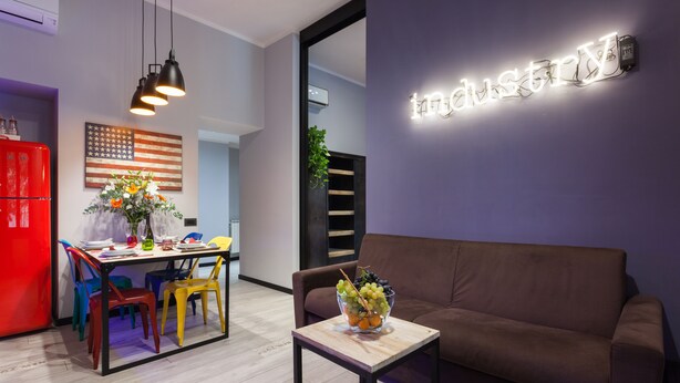 A modern living room with a brown sofa, a small table with a fruit bowl, colorful chairs, a dining area with a red refrigerator, and a wall with a light-up sign. - Three flats close to Vatican - Prati (roma)