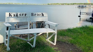 Eine weiße Bank mit der Aufschrift "Kaiser Beach", mit Blick auf einen ruhigen See und einem Regenbogen im Hintergrund.