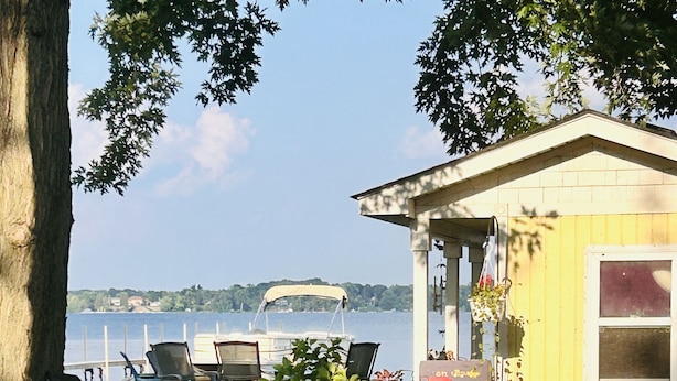 Ein kleines gelbes Häuschen mit Veranda, einem grünen Rasen und Blick auf einen See mit einem Boot im Hintergrund.