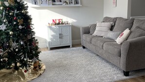Ett vardagsrum med en julgran, en grå soffa och ett vitt skåp med inramade fotografier.