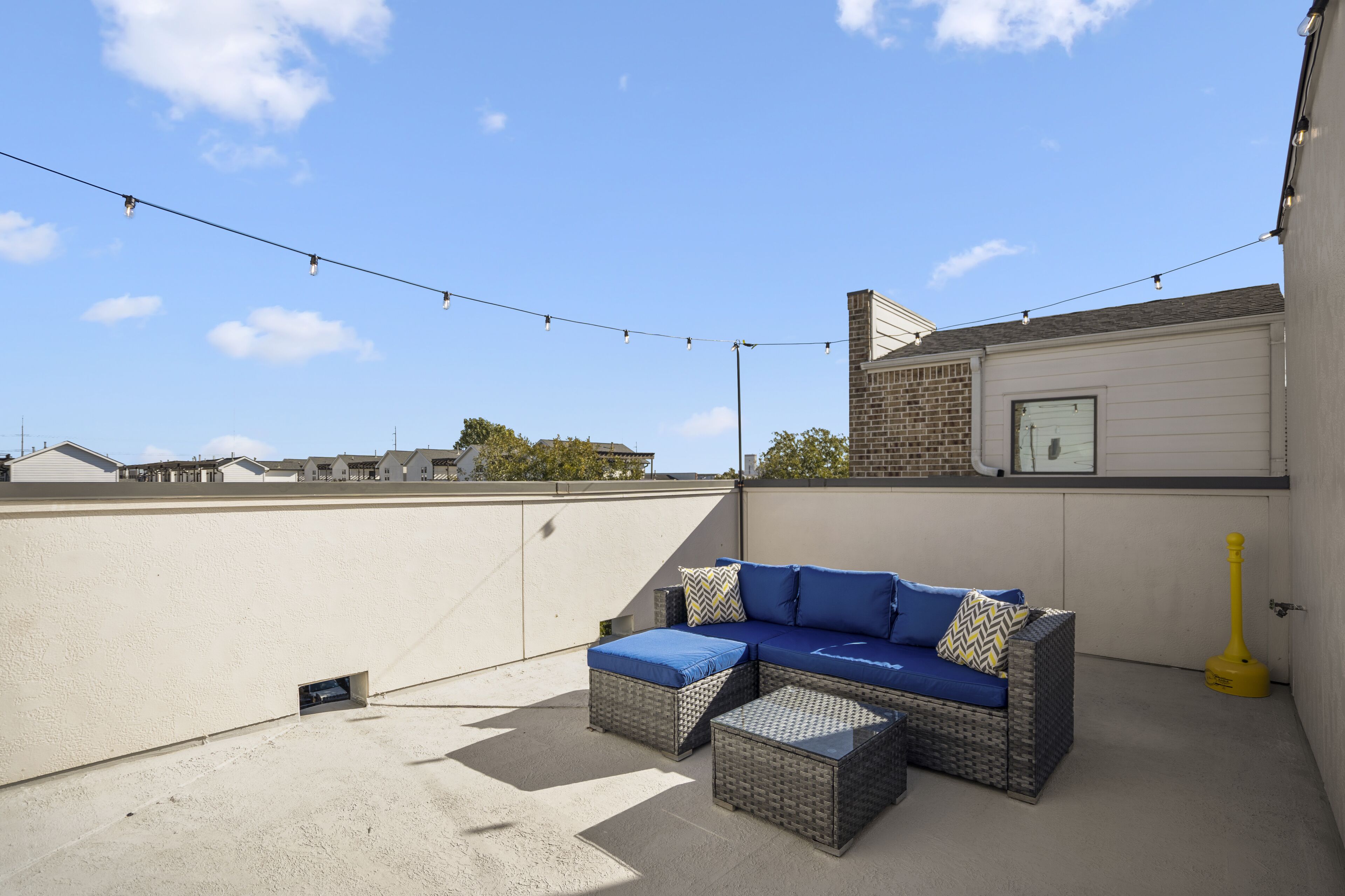 A rooftop patio with a blue sofa, wicker ottoman, and string lights.