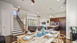 A spacious dining area with a long table set for six, a staircase leading to an upper level, and a modern kitchen with dark wooden cabinets. - Stoic Sanctuary @ East Downtown (Houston)