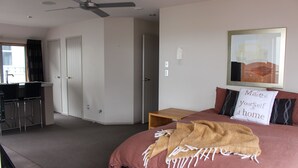 Un dormitorio con cama, escritorio y silla. Hay un ventilador de techo, un cuadro en la pared y un cojín con texto.