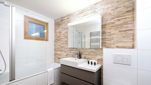 A modern bathroom with a glass shower enclosure, a wooden-framed window, a stone mosaic wall, a white sink with a mirror, and a towel rack. - Chalet Amerhone - Luxury Chalet With Jacuzzi (Riddes)