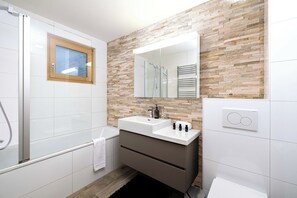 Une salle de bain moderne avec une douche à parois de verre, une fenêtre à cadre en bois, un mur recouvert d’une mosaïque de pierre, un lavabo blanc avec un miroir et un porte-serviettes.