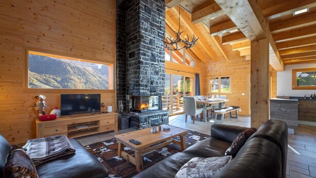 A cozy cabin interior with a stone fireplace, wooden furniture, and a large window overlooking a mountainous landscape. - Chalet Amerhone - Luxury Chalet With Jacuzzi (Riddes)