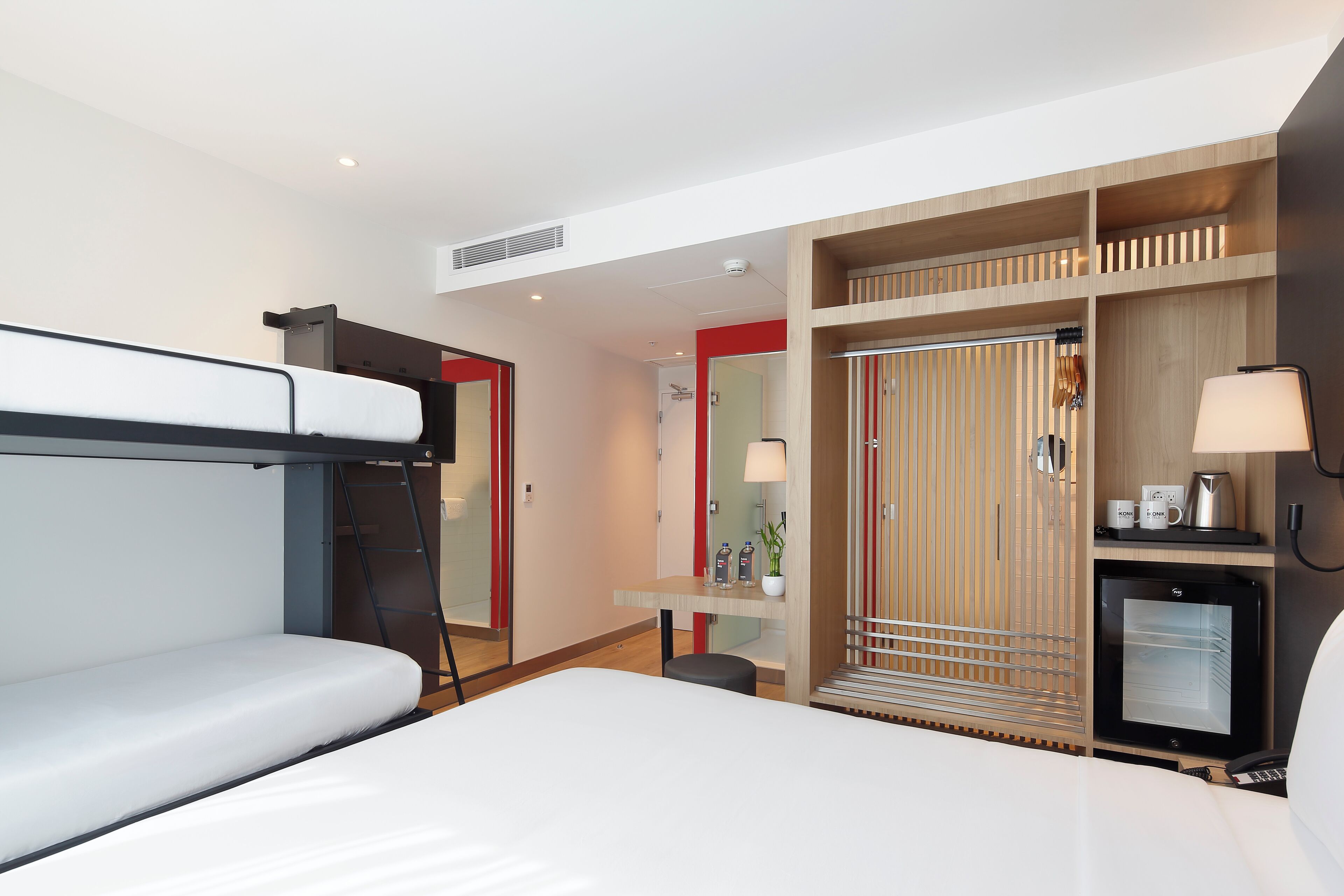 A hotel room with a bunk bed, a desk with a lamp, a small refrigerator, and a wardrobe.