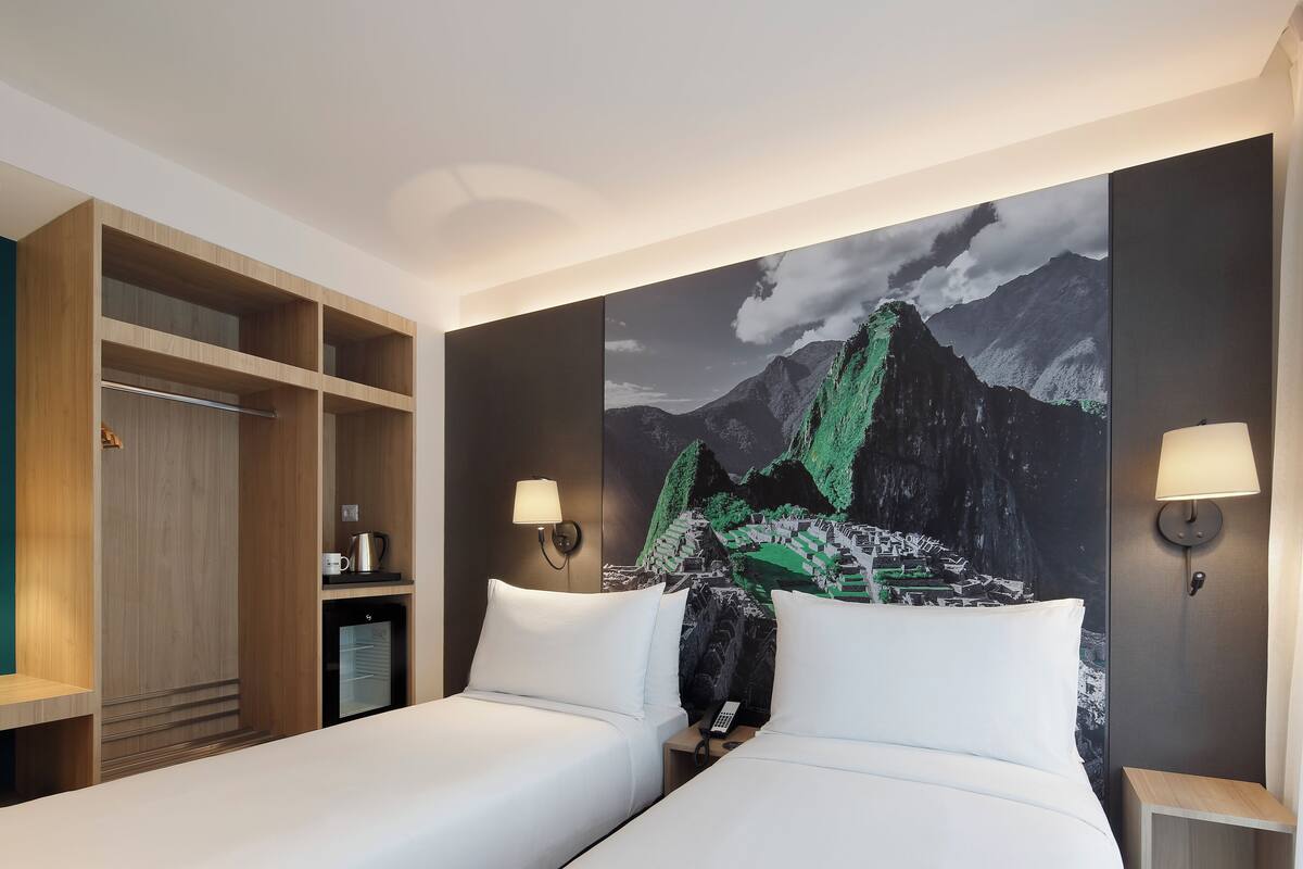 A hotel room with two beds, a wardrobe, a desk with a lamp, and a large mountain artwork on the wall.