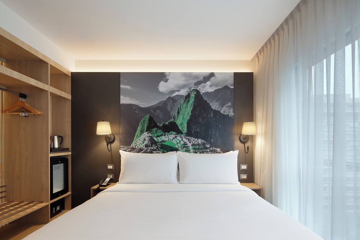 A hotel room with a large bed, two bedside lamps, a flat-screen TV, a wooden wardrobe, and a mountain artwork on the wall.