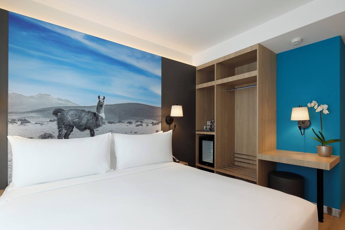 A hotel room with a large bed, a wardrobe, a desk with a lamp, and a wall-mounted TV.