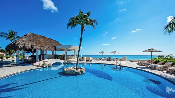 Ein Swimmingpool mit Liegestühlen und Sonnenschirmen, umgeben von Palmen und wolkenlosem, blauem Himmel.