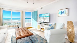 Ein Wohnzimmer mit Meerblick, einem großen Flachbildfernseher, einem weißen Sofa, einer braunen Lederbank und einer gewobenen Stehlampe.