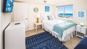 Ein Schlafzimmer mit Bett, Fernseher, Nachttisch, Lampe, einem Fenster mit Blick auf den Strand und einer kleinen weißen Kommode.