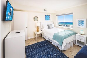 A bedroom with a bed, a TV, a bedside table, a lamp, a window with a beach view, and a small white cabinet.