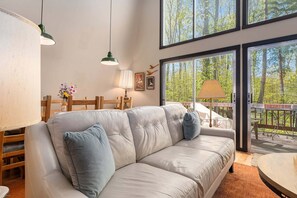 A living room with a sofa, a dining table, and chairs. There are large windows with a view of trees. - 🏡 Sugarloaf Chalet – Pet Friendly Escape | Hot Tub • Fireplace • Loft • Bar Area ➠ 2510 (Cedar)