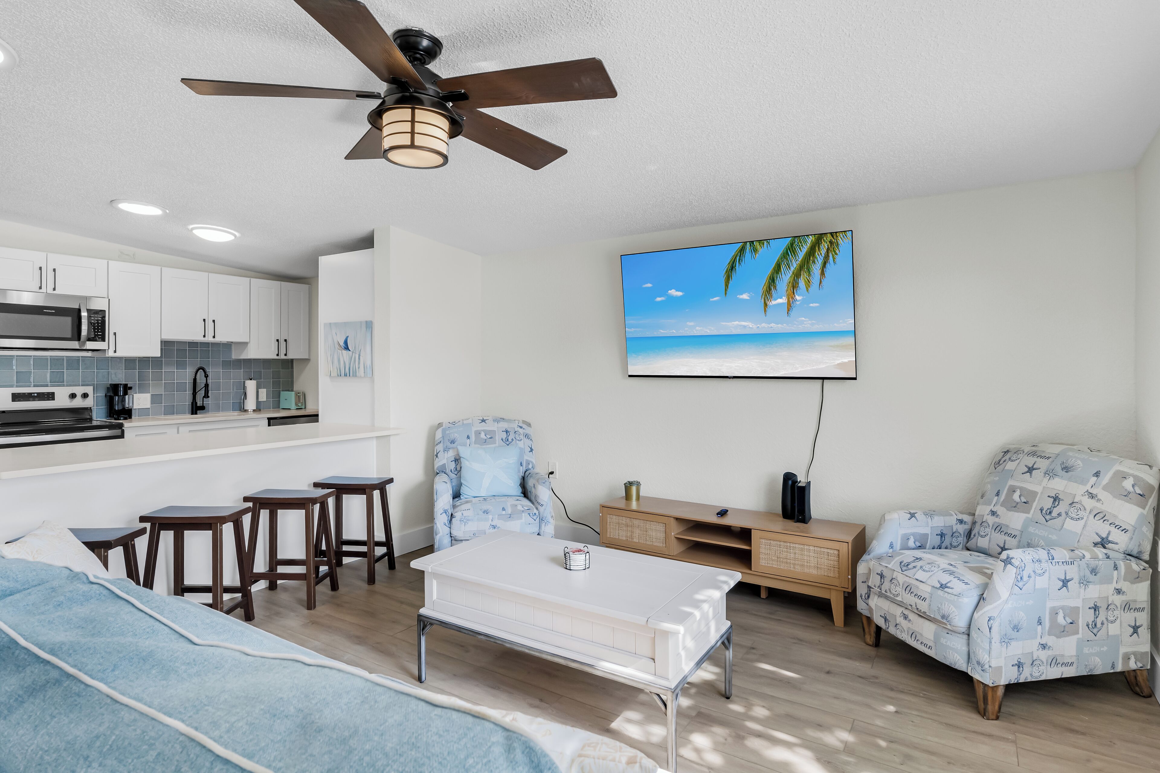 A modern living room with a sofa, coffee table, and two patterned armchairs. A ceiling fan, kitchen area with white cabinets, and a large TV on the wall are also visible.