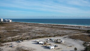 Luftaufnahme eines Parkplatzes direkt am Strand mit Wohnmobilen und Anhängern, einer Straße parallel zum Strand und Gebäuden im Hintergrund.