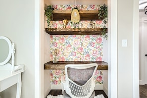 A room with a white chair, a wooden shelf with decorative items, and a floral wallpaper. - Denver Escape| Hot tub+Fire Pit+Games Backyard Fun (Denver)