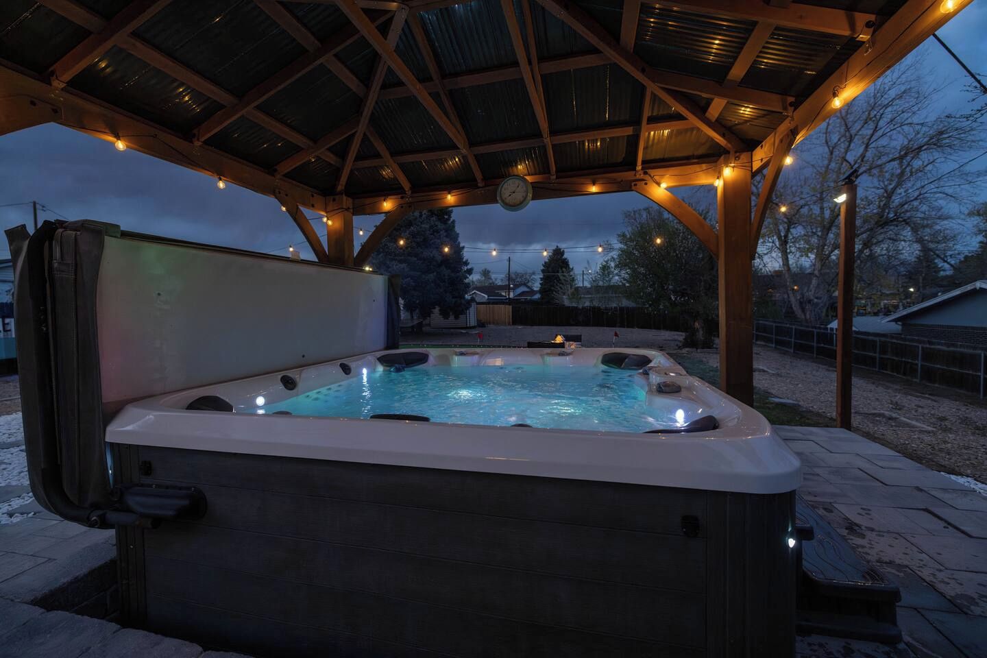 A hot tub under a wooden pergola with string lights, surrounded by a paved area.