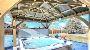 Un jacuzzi bajo una estructura metálica con un reloj, rodeado por una terraza de madera y un patio cubierto de nieve.