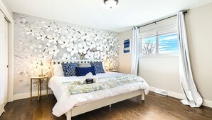 Un dormitorio con cama blanca, un detalle decorativo en la pared, una mesita de noche pequeña y una ventana con cortinas.