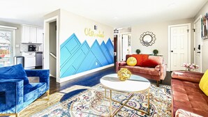 Una sala de estar con decoración azul y blanca, una cocina al fondo y un mural decorativo en la pared.